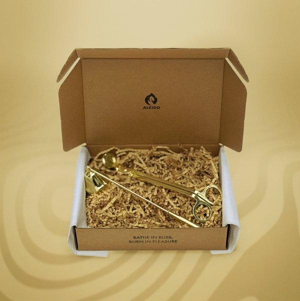 ALKIDO candle care set in kraft box featuring gold wick trimmer and snuffer, nestled in shredded paper, with ‘Bathe in Bliss, Burn in Pleasure’ printed inside lid, on a soft yellow background.
